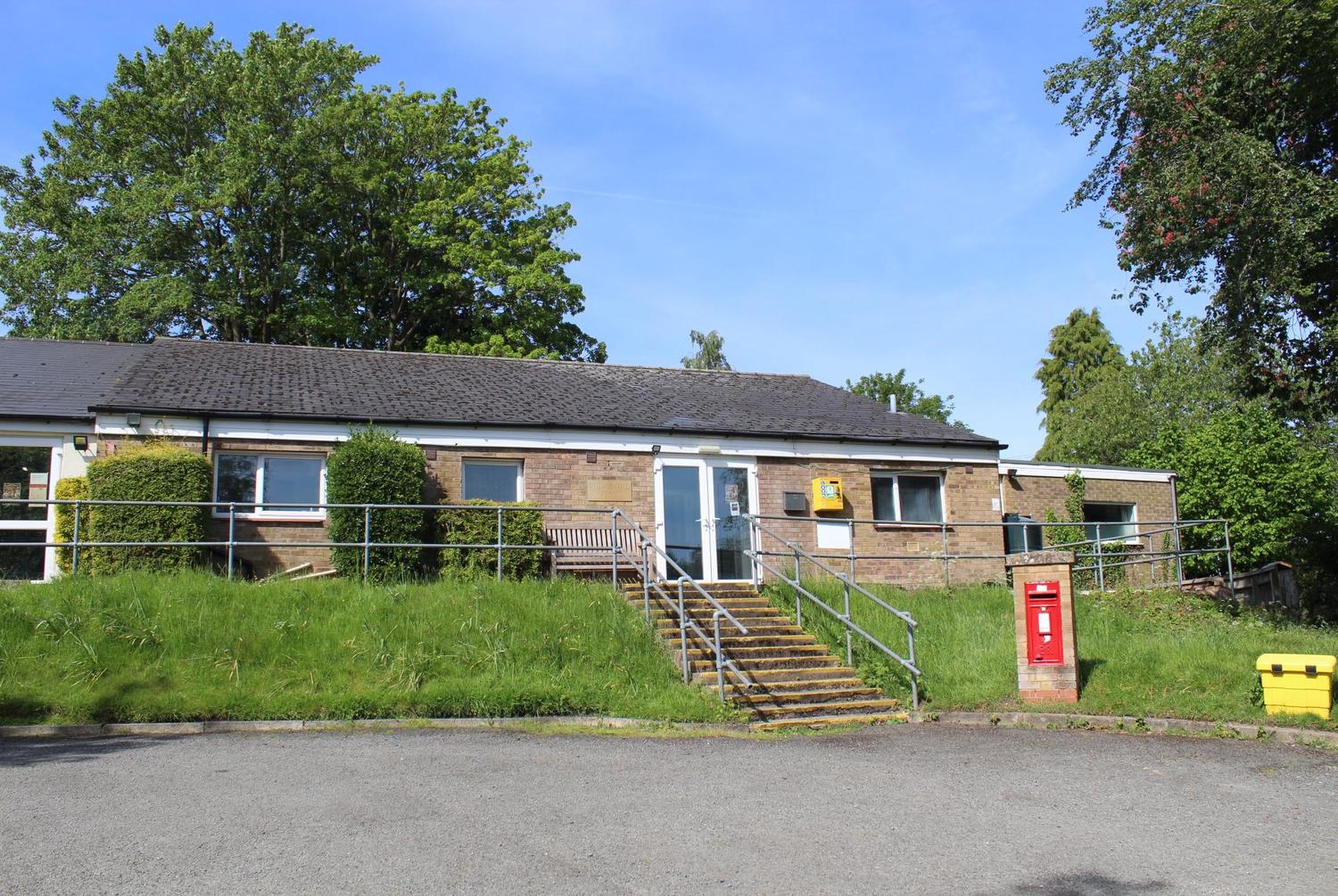 Lighthorne village hall building with steps and railings, surrounded by grass and trees, under a cle